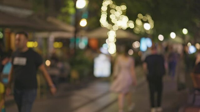 People walk along pedestrian street of big city. Evening, illumination, cafes and restaurants. Summer atmosphere of the city. Defocused shot