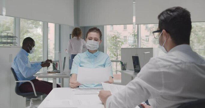 Female employer wearing face mask holding cv listening male candidate at job interview