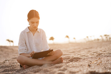 Naklejka na ściany i meble Cut teenager girl using tablet on the background of the beach during sunset. Summer vacation concept, studying online with tablet