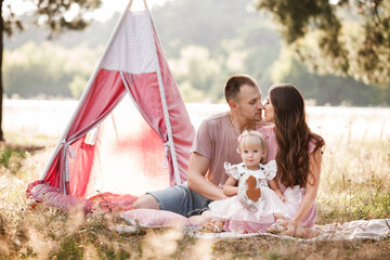 Mom, dad and little daughter are sitting next to wigwam decoration in the park. Family spending time outdoor in summer, having fun together. Girl are dressed in pink dress © Andriy Medvediuk