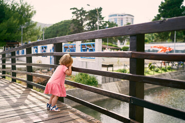 Little girl stands on the bridge, leaning on the fence