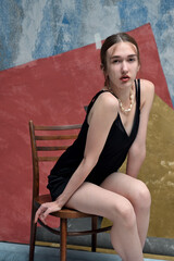 A lovely serious girl in a black dress sits on a chair in the studio.