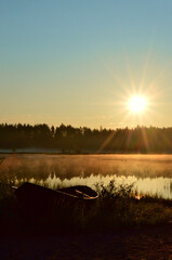 Late summer sunrise on a small pond. A mist floats above the water. The rising sun gilds the landscape with its light. There is an old boat on the beach.