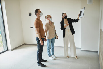 Full length portrait of female real estate agent wearing mask while giving apartment tour to young couple, copy space