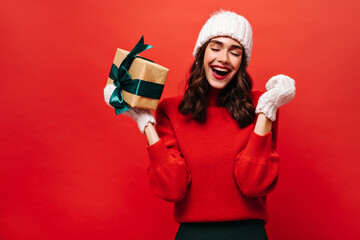 Portrait of excited curly girl in white hat and knitted mittens rejoices and holds gift box on isolated red background.