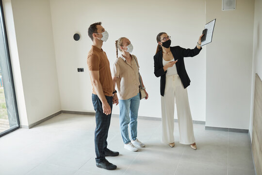 Full Length Portrait Of Female Real Estate Agent Wearing Mask While Giving Apartment Tour To Young Couple, Copy Space