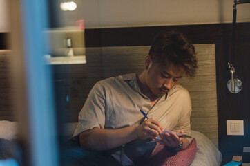 Young relaxed asian man traveller using smartphone, computer and taking notes in hotel room while on vacation