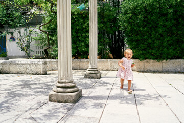 Fototapeta na wymiar Little girl walks along a tiled path near the columns against the background of green magnolia bushes