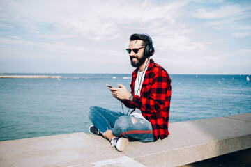 Cheerful hipster guy in trendy apparel sitting outdoors at coastline and laughing while listening positive audio book using electronic headphones for noise cancellation and cellphone knowledge app