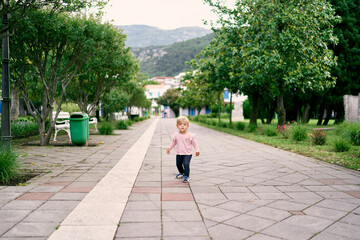 Fototapeta na wymiar Little girl walking along the paved road in the park against the background of green trees and mountains