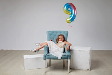 Happy preteen girl with long hair in festive dress sitting carefree on armchair over white pastel...