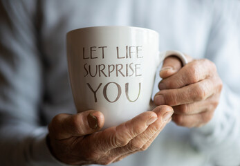 Hands of old man holding cup mug coffee tea with inspirational quote Let Life Surprise Yoy