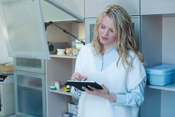 Beautiful blonde medical resident keeps statistics of pharmacological drugs in the doctor's office, writing the data on the tablet with a pen.