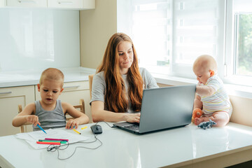 Young focused business mom with two children working remotely on a laptop at home in the kitchen.