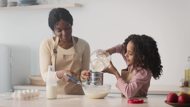 Family bakery. Little african american girl helping her mother with preparing dough, adding flour to sifter at kitchen