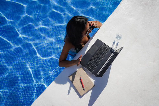 remote online working digital nomad woman calling with mobile phone & laptop on a white table in a sunny blue water pool on Workation
