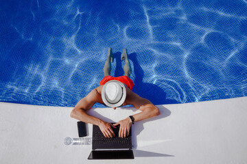 bird view of remote online working digital nomad man on workation with hat &  laptop at a white table standing in a sunny turquoise water pool