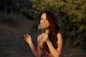Beautiful happy woman blowing big dandelion. Outdoors, enjoy nature. Healthy smiling woman on summer lawn. Allergy free concept. Gorgeous slim caucasian, free space for text