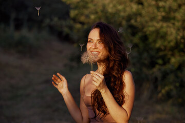 Fototapeta premium Beautiful happy woman blowing big dandelion. Outdoors, enjoy nature. Healthy smiling woman on summer lawn. Allergy free concept. Gorgeous slim caucasian, free space for text