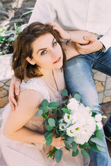 beautiful bride looks at the camera sits near her husband's feet, holding a bouquet