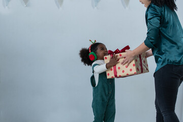 Happy little African kid girl with green bib clothes and earmuffs get gift box from Asian mother in Christmas holiday. White background