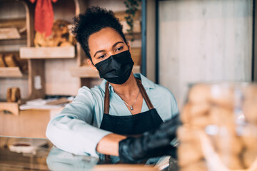 African American middle aged female worker with protective mask on face working in bakery....