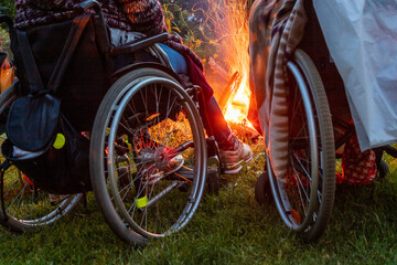 Fototapeta na wymiar Two people in wheelchairs enjoying campfire at night. Low angle view to wheelchairs wheels during summer nighttime at fireplace