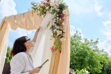 Decorating arch with textiles with flowers and plants. Woman organizer, owner, with digital tablet...