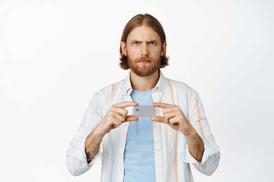 Image of angry serious blond guy, frowning upset, showing credit card and looking at camera concerned, standing in shirt against white background