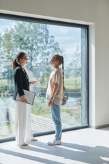 Full length portrait of female real estate agent giving apartment tour to young woman while standing by floor to ceiling window