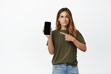 Skeptical, woman with doubts pointing finger at mobile phone empty screen, smirking and frowning unamused, concerned about smth on smartphone display, white background