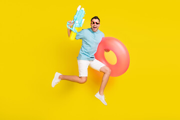 Fototapeta na wymiar Photo of funky sweet young guy dressed blue t-shirt eyewear jumping holding inflatable rings water gun isolated yellow color background