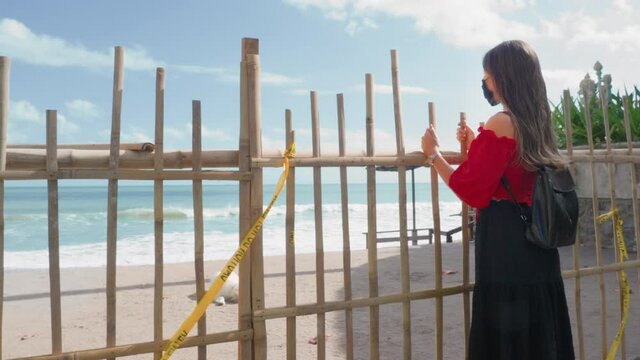 A lonely young girl with long hair, dressed in a red blouse, she stands at the bamboo fence that blocks her way to the paradise beach