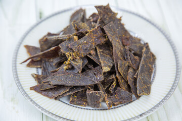 Deer meat (Lat. Cervidae) dried, cut into strips on a round plate with a wooden table background. Food is a delicacy snack for beer.