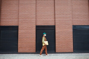 Wide angle shot of delivery man wearing mask while walking by brick wall outdoors, copy space