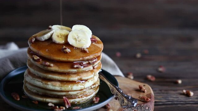 Syrup being poured over a delicious stack of pancakes with fruit and nuts over a rustic table. Selective focus on bananas with blurred foreground and background.