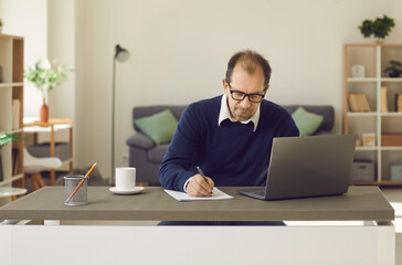 Mature caucasian man in casual clothes sitting at desk using laptop screen writing on paper notebook at home office portrait. Online distant work, remote education or e-learning, training courses