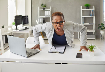 Angry stressed black business woman in glasses leaning over office desk with laptop computer and screaming at you. Crazy frustrated female boss in rage shouting at camera. Concept of stress at work - Powered by Adobe