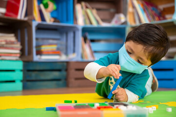 Mexican baby in school with face mask playing with colored pieces on a mat, back to school 