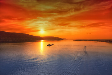 Fiery sunset over the river and mountains, red sky, blue river, boats and cargo ship barge float down the river at sunset.