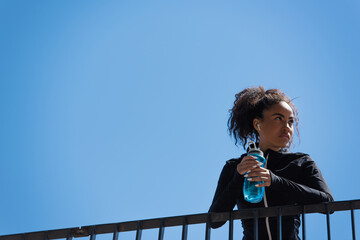 Low angle view of african american sportswoman in earphone holding sports bottle with blue sky at...