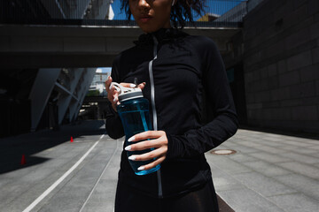Cropped view of african american sportswoman in earphone holding sports bottle outdoors