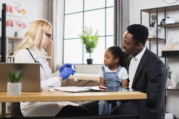 Obraz premium Competent female pediatrician providing medical consultation for african american man with cute little daughter. Father with child visiting caucasian doctor for checkup.