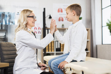 Side view of caucasian boy giving high five to pleasant female doctor at hospital cabinet. Male patient getting professional health treatment at modern clinic.