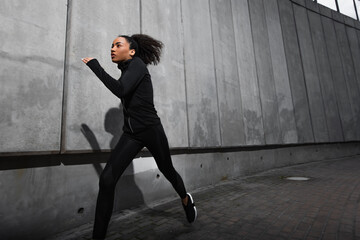 Young african american sportswoman running near concrete wall with shadow on urban street