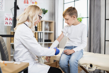 Caucasian female doctor wounding bandage around little boy's arm at modern hospital. Medical worker...