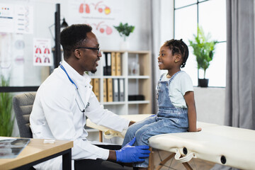 Obraz premium Side view of african american doctor in white lab coat and gloves examining health of little cute girl at modern hospital. Concept of medicine and childhood.