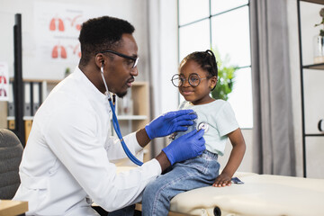 Pediatrics and regular check up concept. Pleasant caring Afro-American man physician checking littel African kid girl's heartbeat and breath using stethoscope, working in medical center
