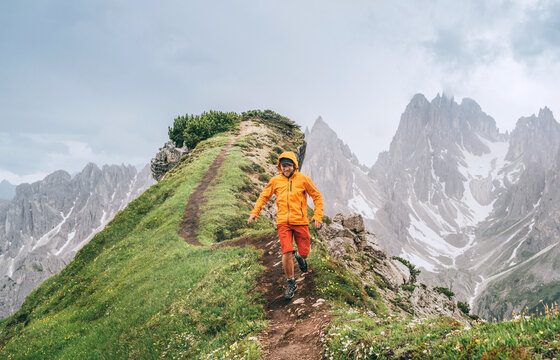 Dressed bright orange soft shell jacket backpacker running  by green mountain path with picturesque Dolomite Alps range background,. Active people and European mountain hiking tourism concept image.
