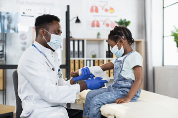 Lovely cute dark-skinned small girl in facial mask sitting on couch in medical office, while...